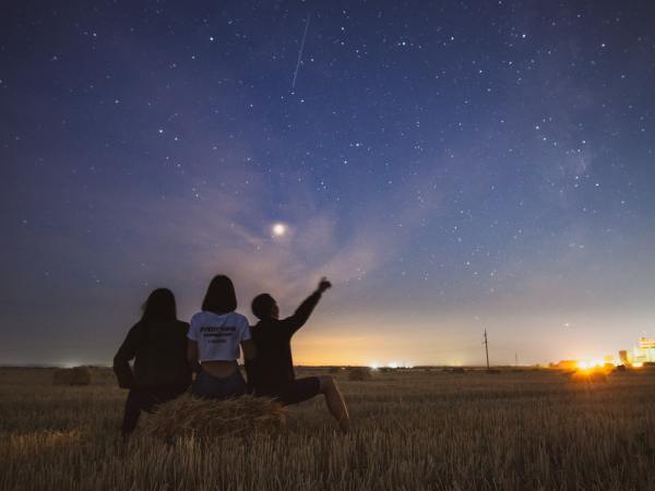 Three people watching the night sky while sitting on a hay bale in a field