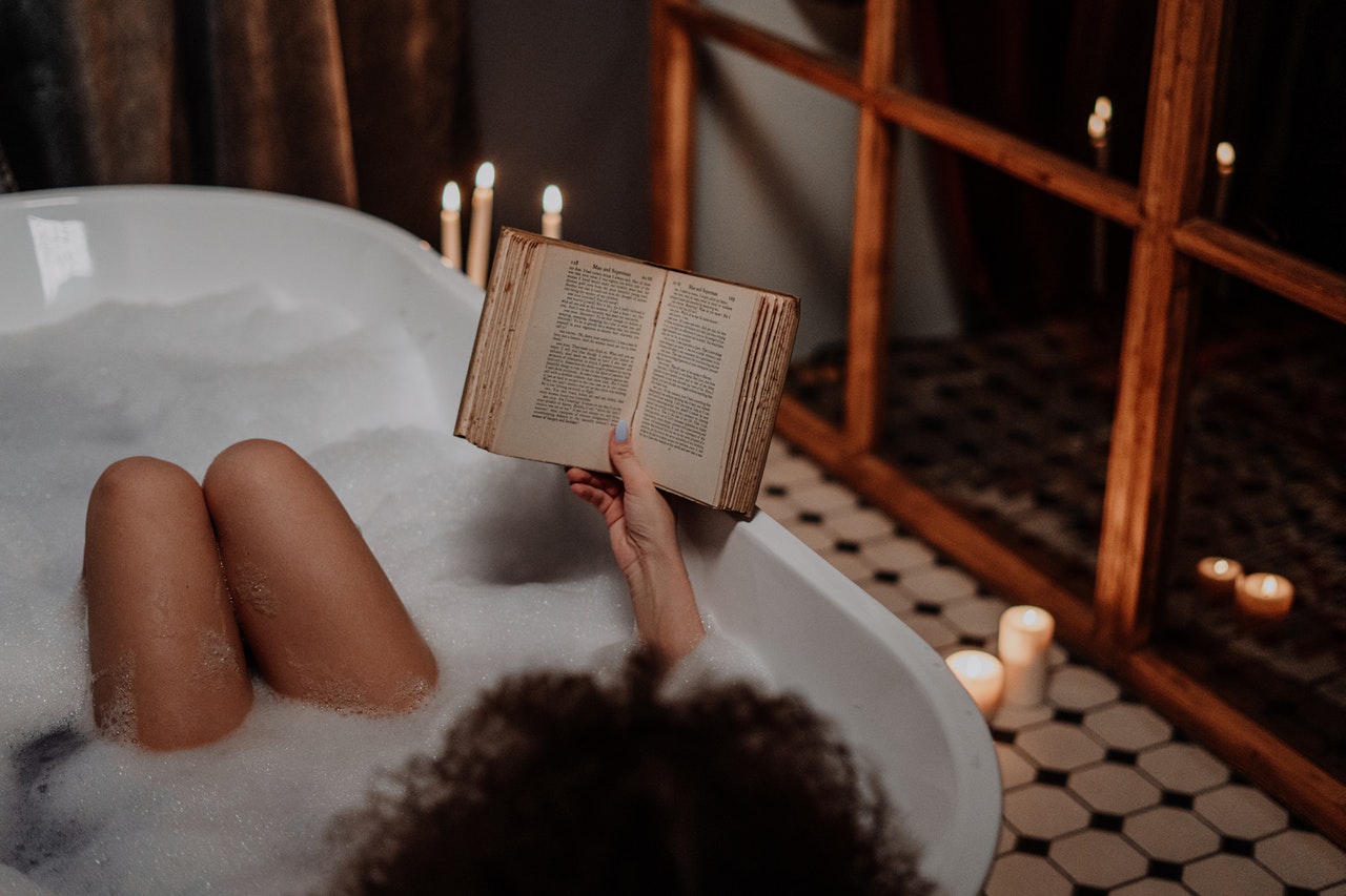 a woman reads an old book in a bath tub full of bubble bath surrounded by candles