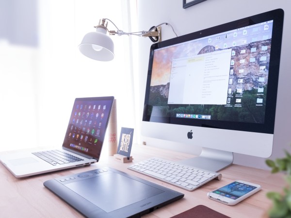 An Apple macOS laptop, desktop computer, tablet, and iOS iPhone on a wooden desk.