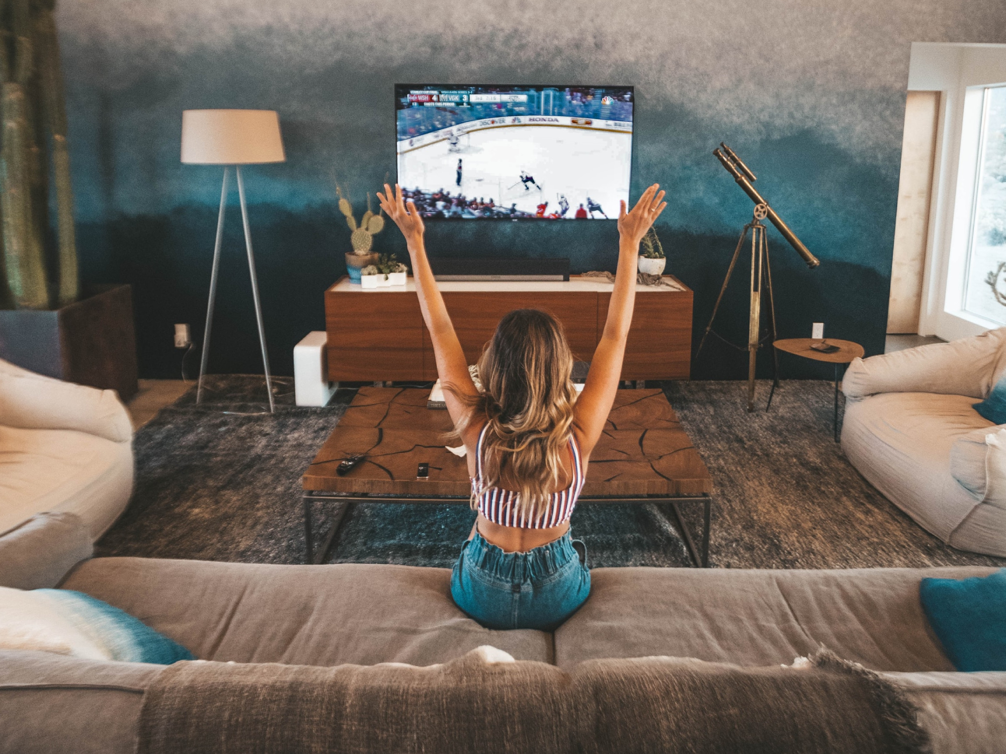 A woman cheering while sitting on a brown couch and watching a hockey game on a large flat-screen TV