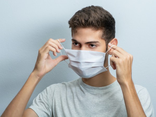 A man in a gray t-shirt removing or putting on a surgical face mask by only touching the ear loops.