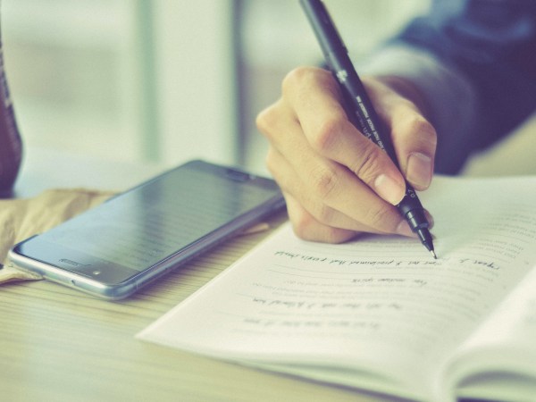 A person using a pen to write in a paper notebook, with a phone on the table next to them for storing digital notes.