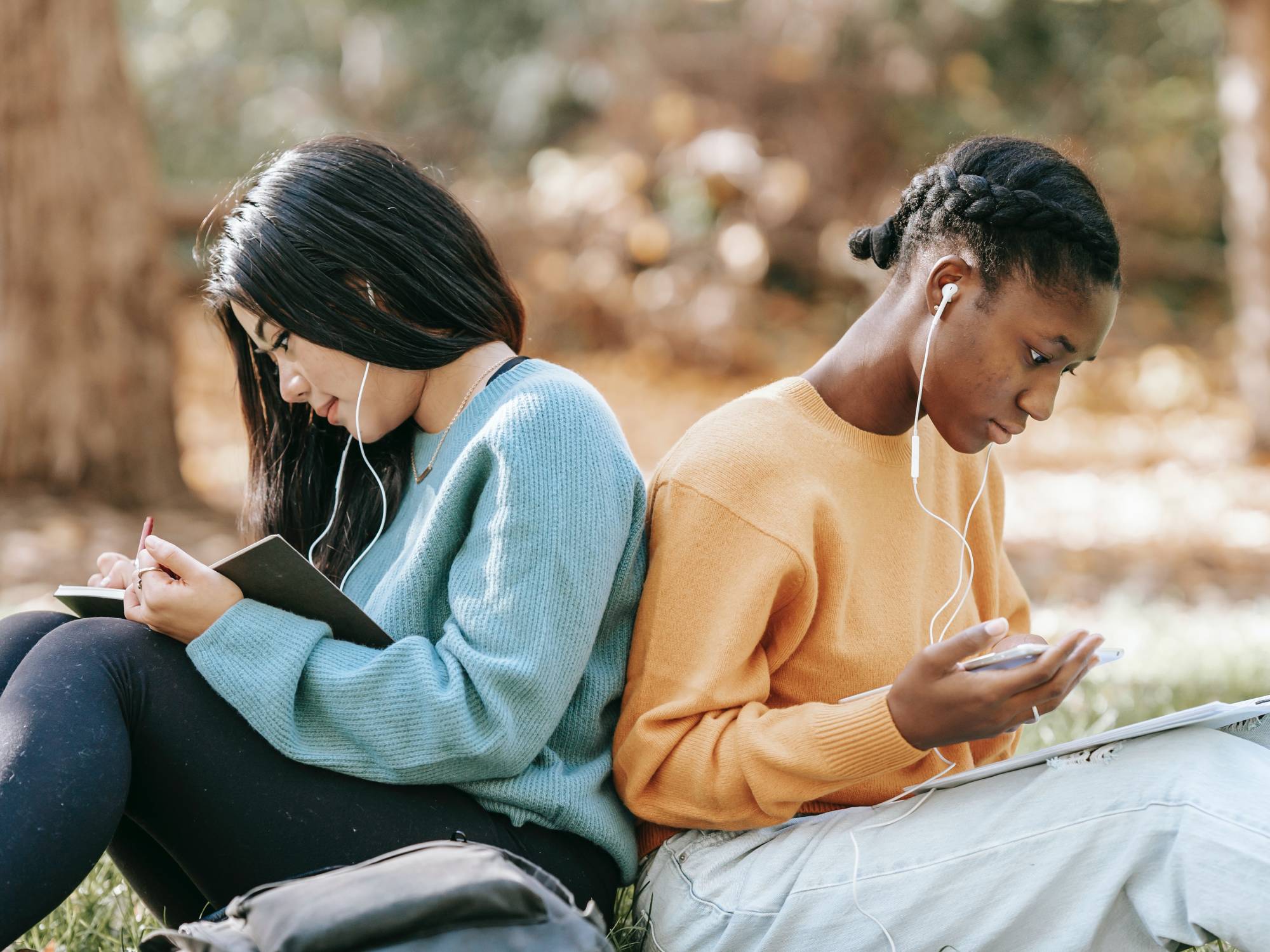 Young people back to back, sitting in a park, studying with their phones.