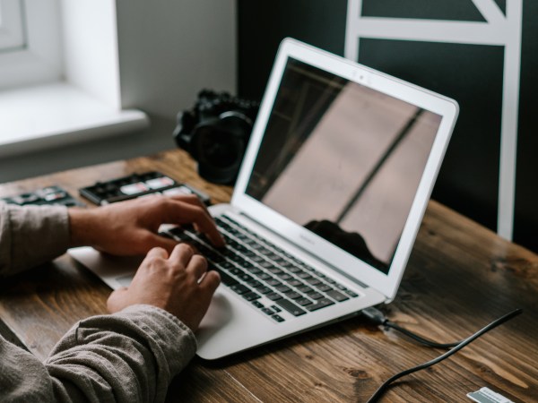 Laptop on a wooden desk near a window with a person's hands on the keyboard.