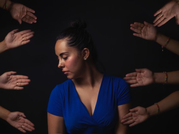 A woman in a blue dress looking away and down to the side, as eight hands reach out to her from the edge of the frame.