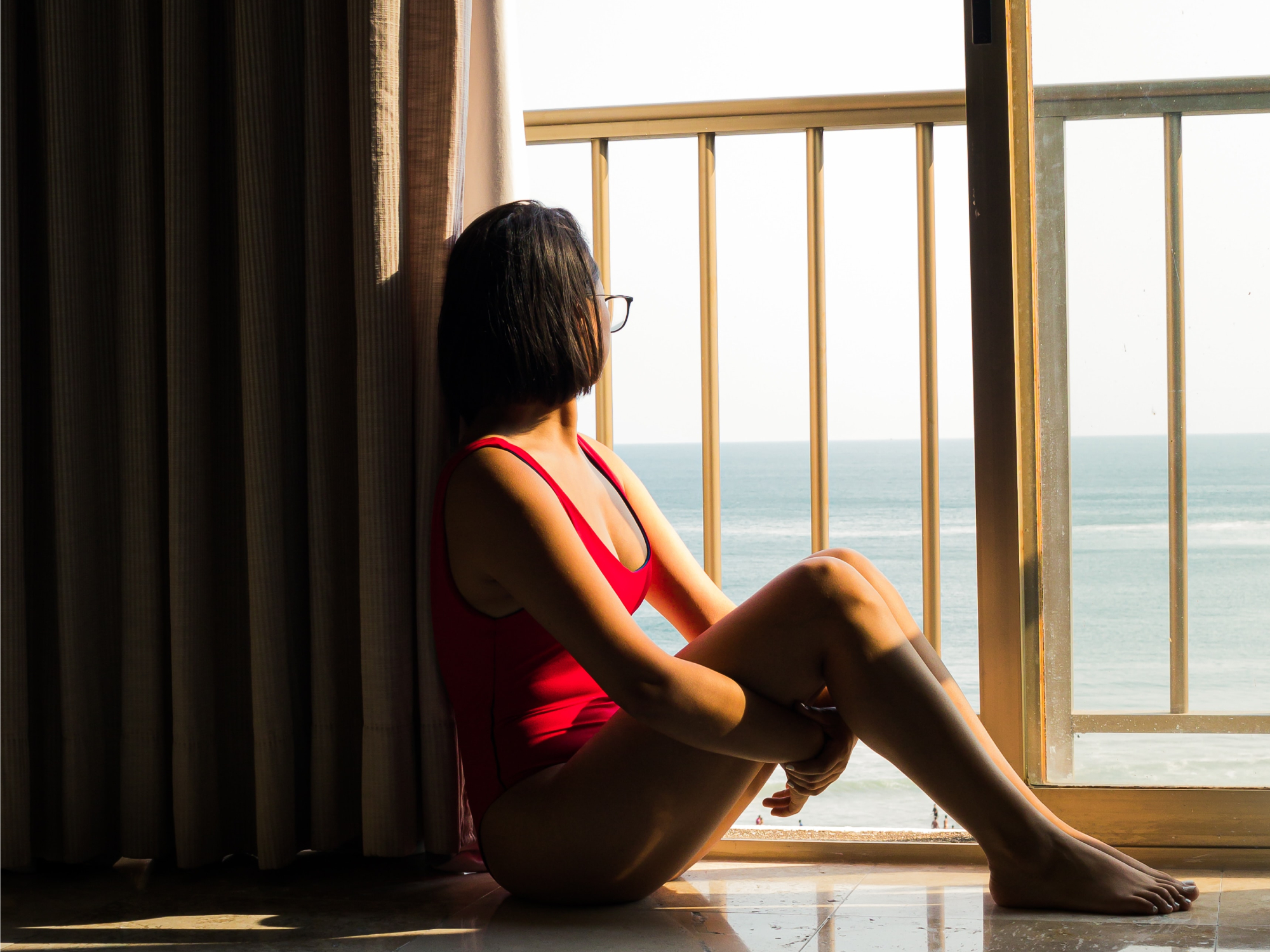 A woman in a red bathing suit sitting on the floor by a sliding glass door, staring at the ocean outside.