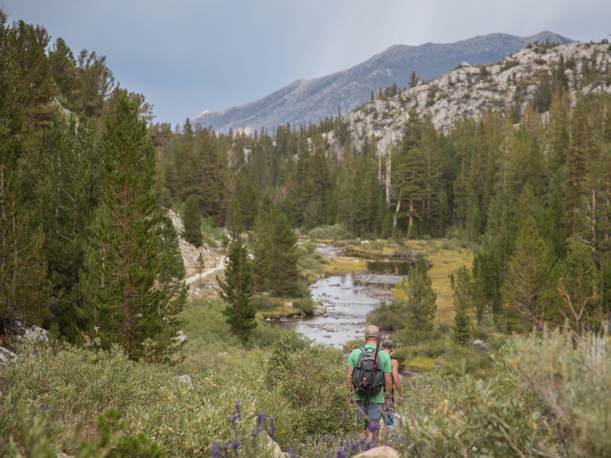 Two people walking on a trail in the wilderness, heading toward a body of water and some mountains