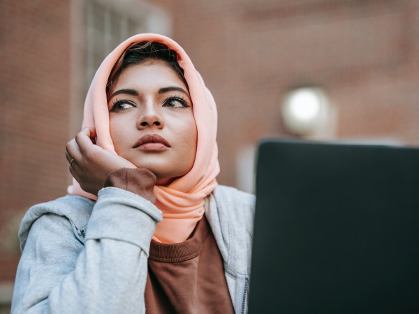 A Muslim woman daydreaming while sitting in front of a laptop outdoors.