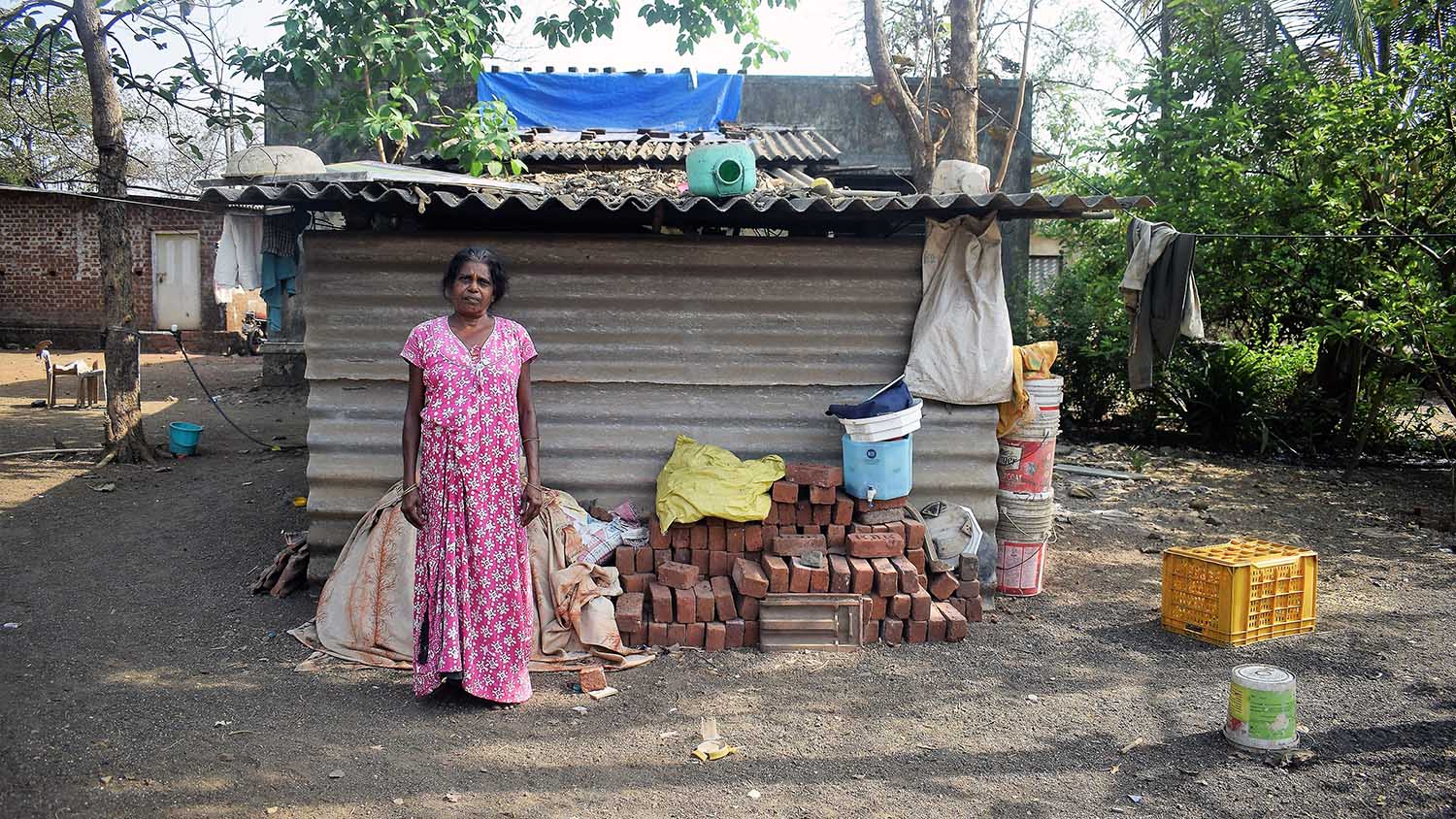 Domestic worker in a pink dress standing in front of a tin home and pile of bricks