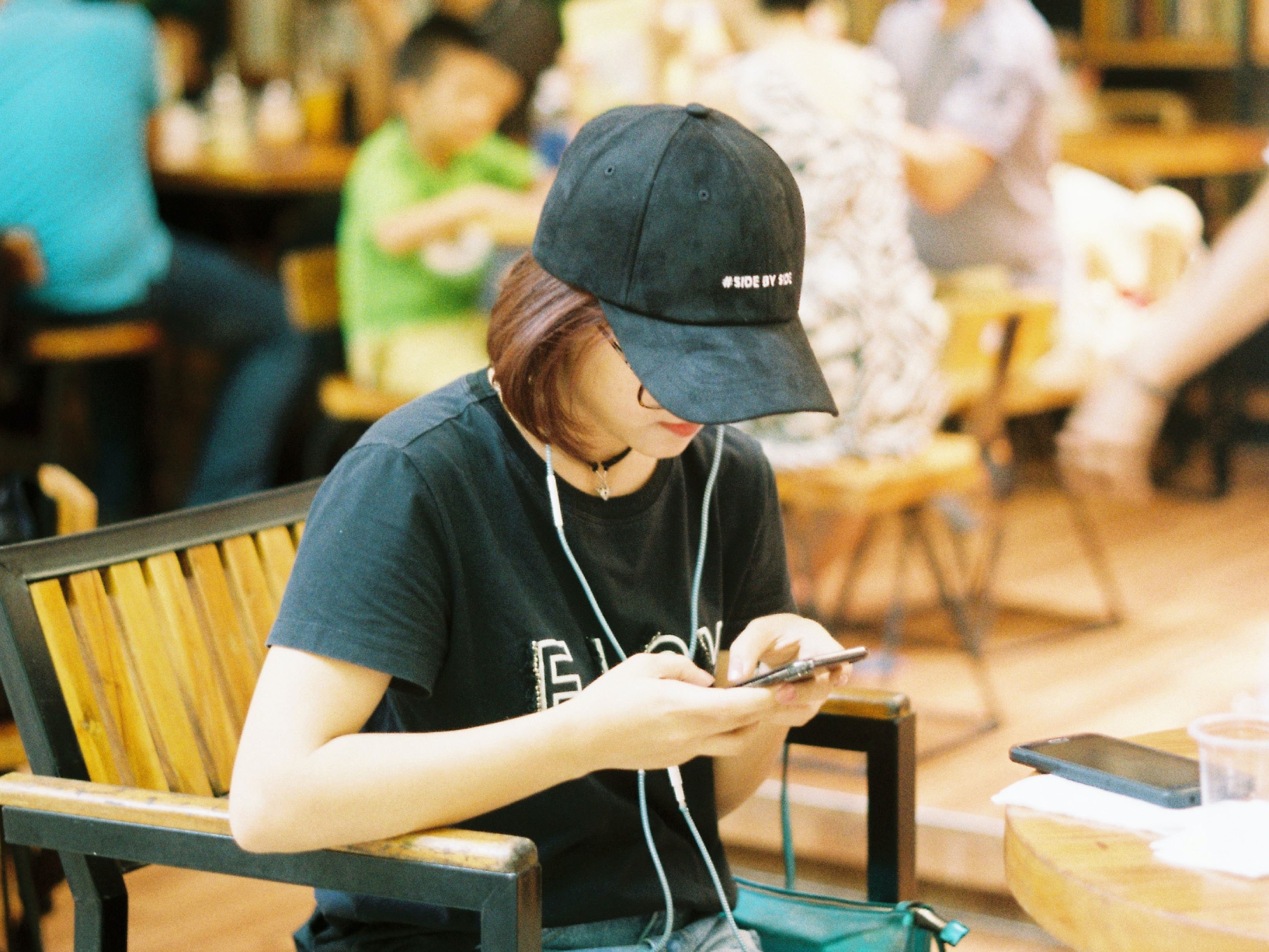 A person sitting at a coffee shop, using a phone while wearing headphones and a cap.