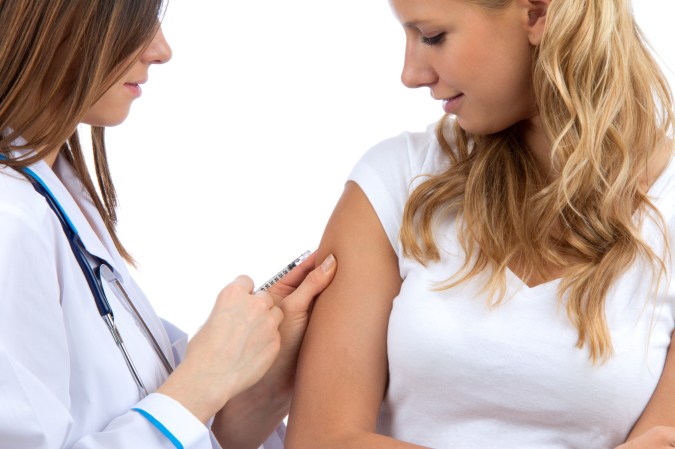 Person with long blond hair getting a shot from a doctor with a stethoscope