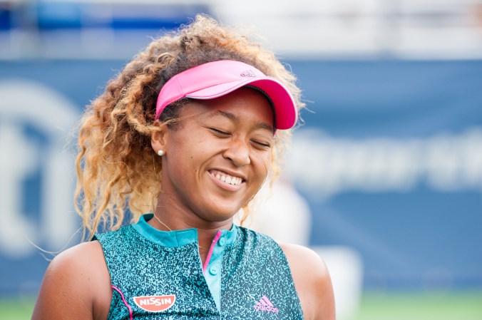 Naomi Osaka smiling on the tennis court in a pink visor