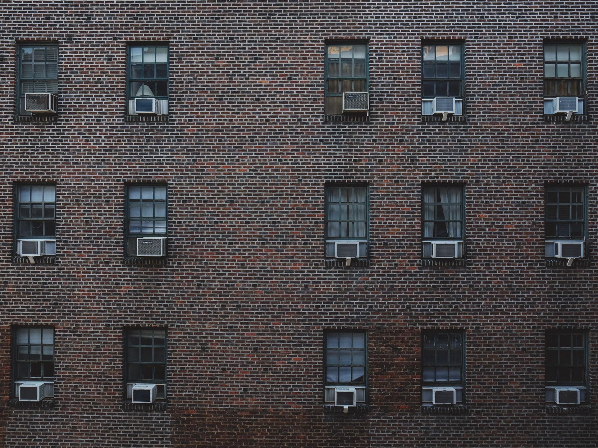 One side of a reddish-brown brick building, with 15 windows arranged in a grid on the façade, each containing a separate window air conditioning unit.