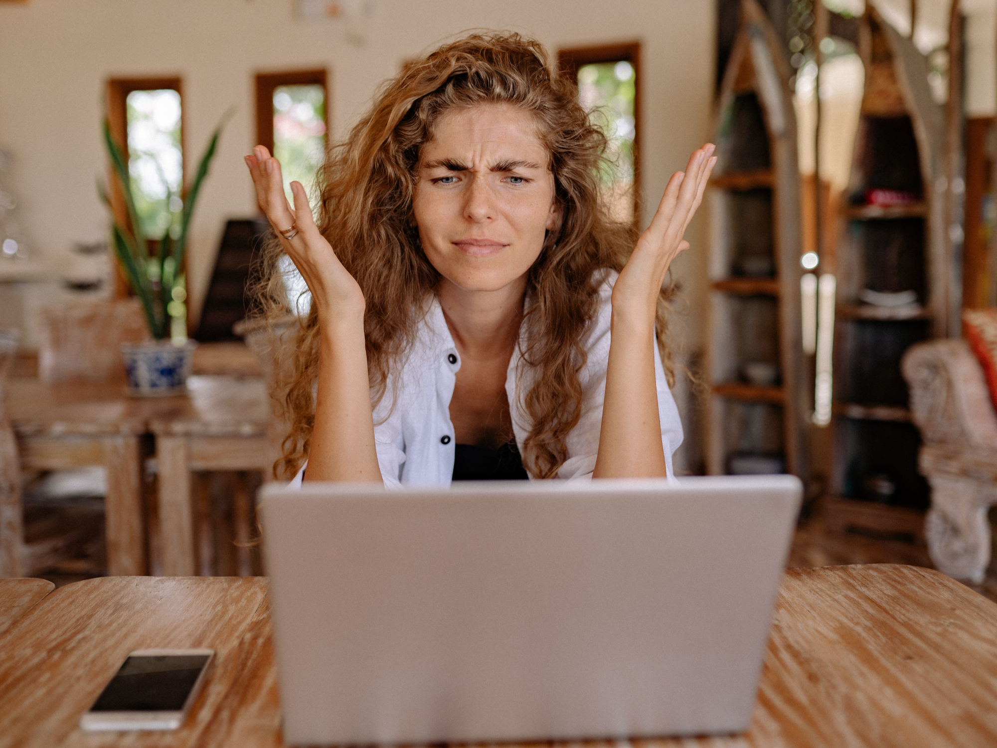 A woman with curly, dirty blonde hair sitting in front of a gray laptop with her hands in the air in an act of confusion or frustration.