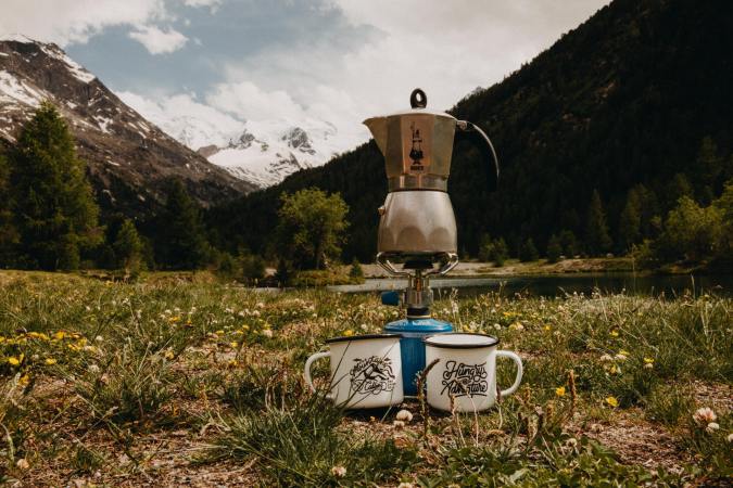 Moka pot on a burner in front of a snowy mountain with two enamel cups