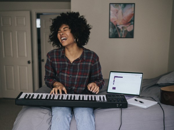 A person sitting on a bed, enjoying playing a keyboard that's hooked up to a laptop.