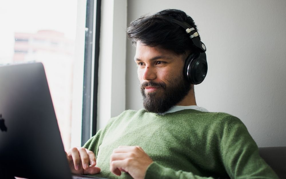 A young man with dark hair and beard and a pair of black headphones placed in his ear and a laptop in front of him.