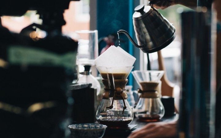 A person who is filling a cup of coffee with white pizza placed on top and a silver kettle with coffee in it.