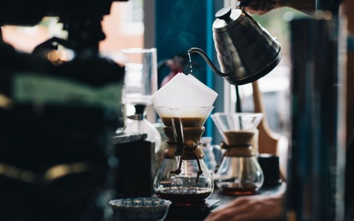 A person who is filling a cup of coffee with white pizza placed on top and a silver kettle with coffee in it.