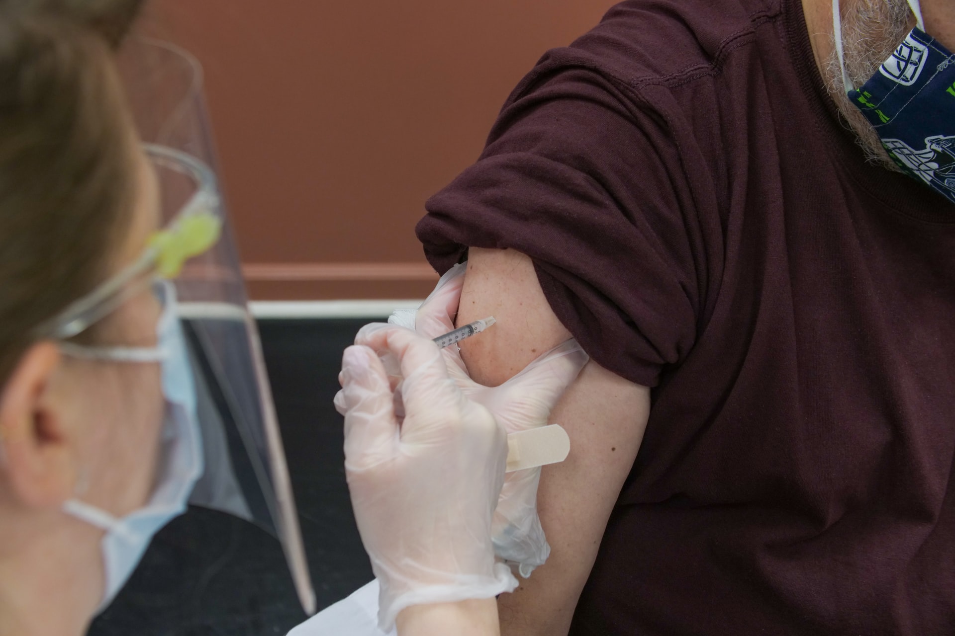 A healthcare professional administers a shot in an arm to a person in a brown shirt.