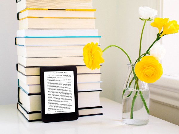 A Kobo e-reader leaning against a stack of books next to a glass jar with yellow flowers in it.