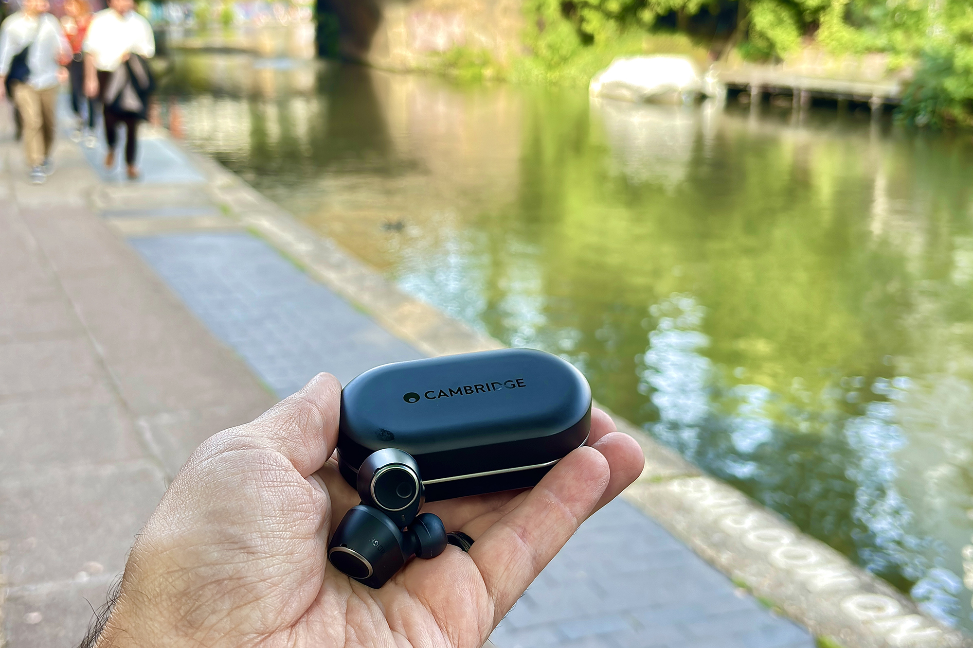 Black Cambridge Audio M100 TWS ANC earbuds held in a hand on the side of the Regent's Canal Towpath in London