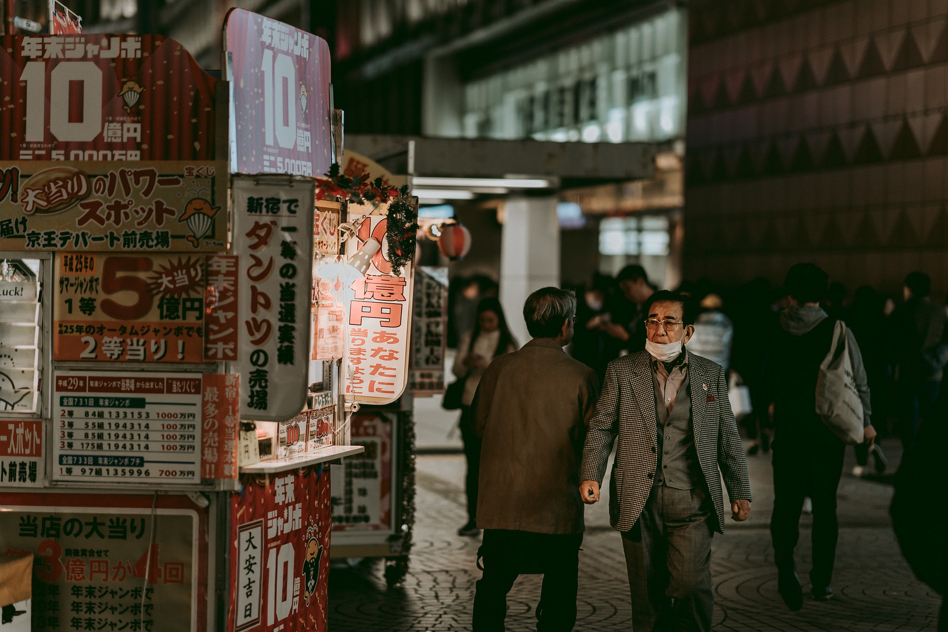 A man wearing a grey suit and glasses has a white surgical mask partially covering his mouth and chin. He is walking down a cobblestone street facing towards the camera while others walk facing the other way. It is nighttime and a building and street vendor are in the background.