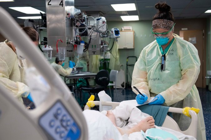 A nurse in a mask and gown writes in a medical chart in front of a patient in a hospital bed.