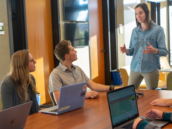 A person speaking to a group of people who are sitting at a wooden table with laptops in front of them.