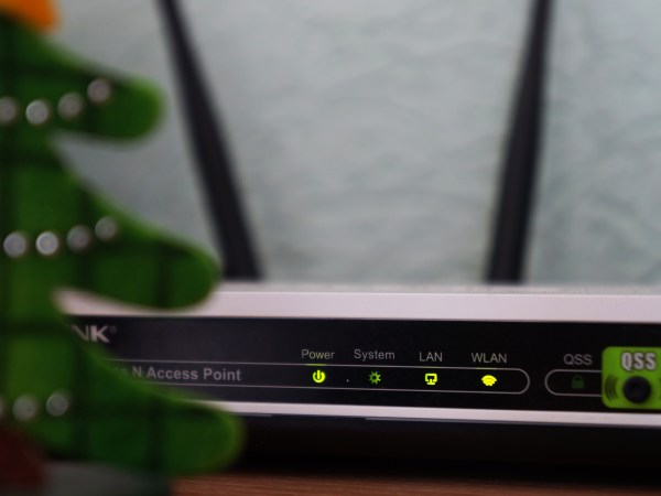 A WiFi router on a table against a gray wall, behind a plant.