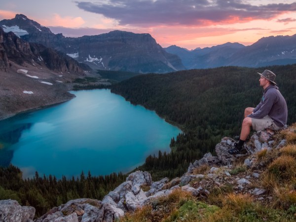 A person wearing a wide-brimmed hat sitting on some rocks high above a bright blue glacial lake in the mountians.