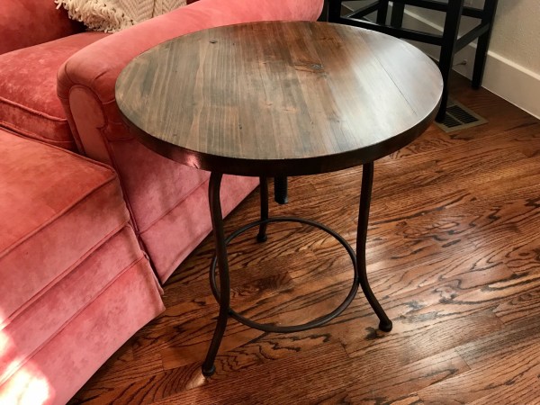 A refinished wooden side table with black iron legs, on a hardwood floor next to a pink armchair.