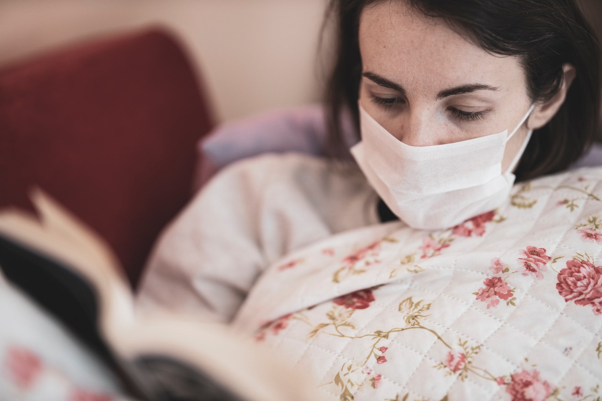 A woman in bed wears a mask and reads a book.