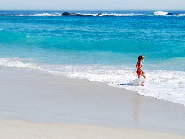 A young child standing alone at the edge of a bright blue ocean, playing in the foam.