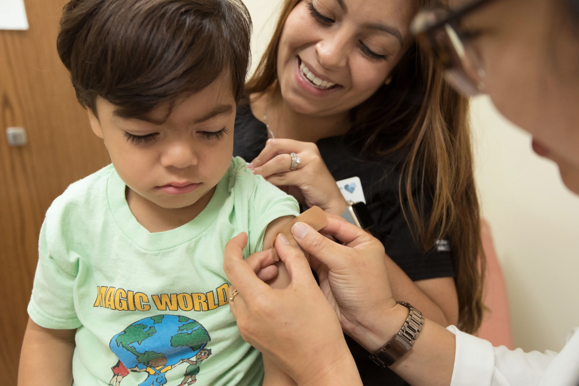 A small child is having a bandage put on his arm after receiving a shot. A caregiver stands behind him.