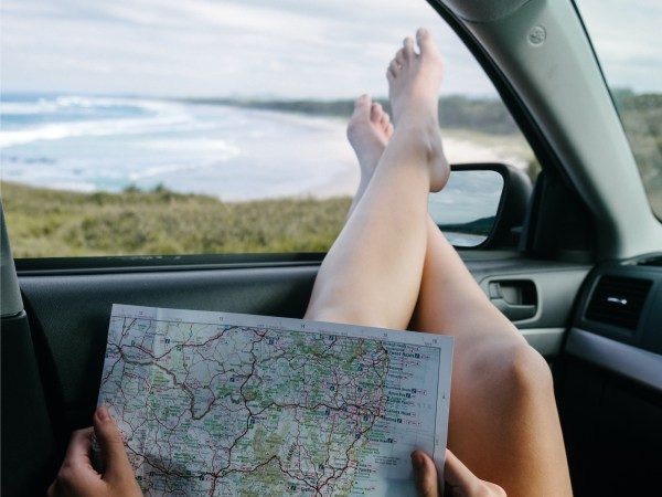 A person sitting in a car with their bare feet out the open window, looking at a road map while overlooking an ocean.