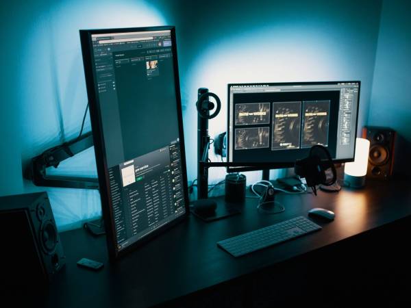 A black desk with a vertical monitor setup, in addition to a horizontal monitor, with both screens backlit with white light.