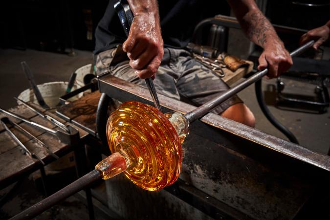 a human's hands hold a metal rod as they manipulate a rounded piece of orange glass