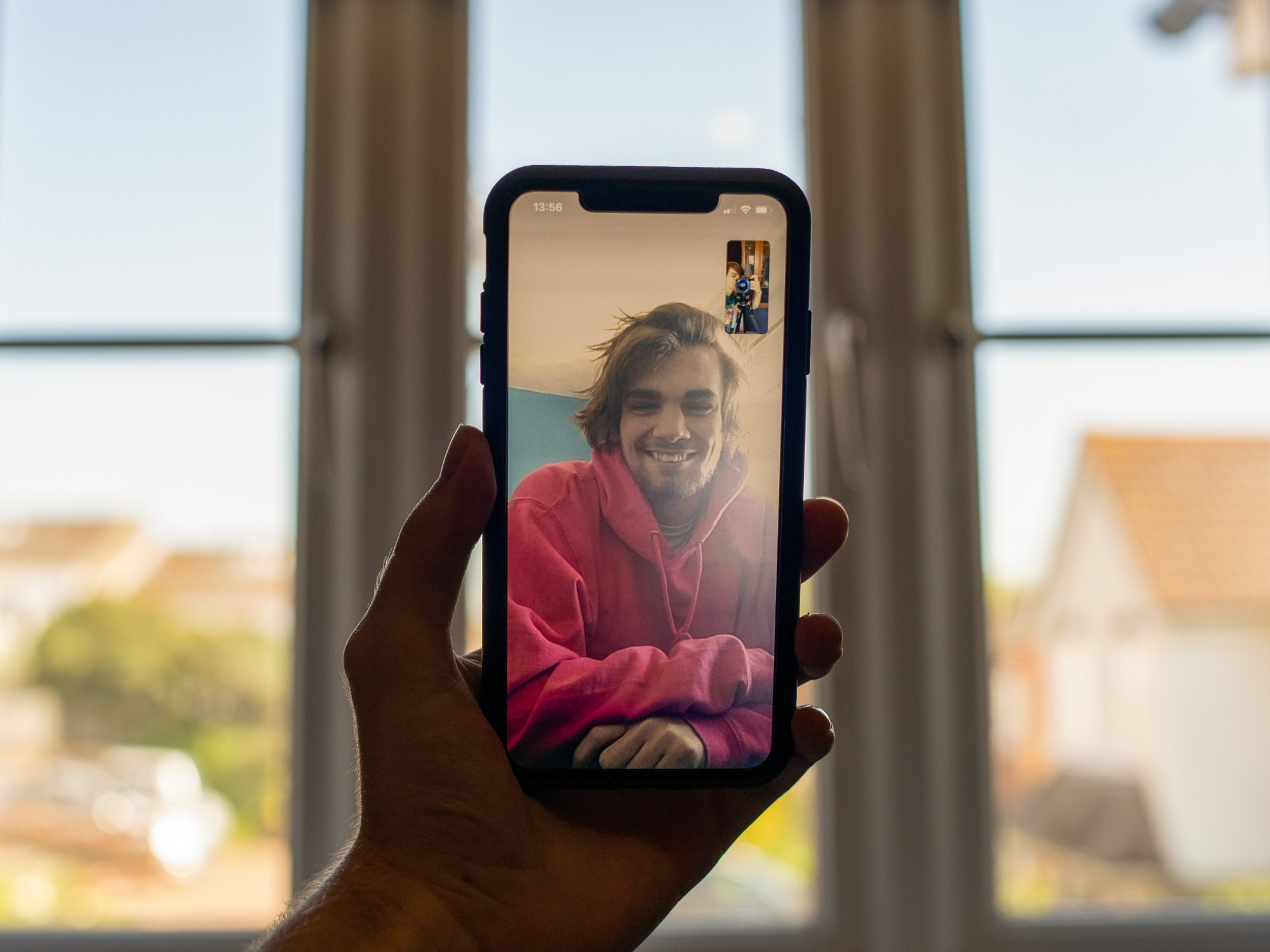 A person holding an iPhone in front of a window while having a FaceTime call with someone in a red sweatshirt.