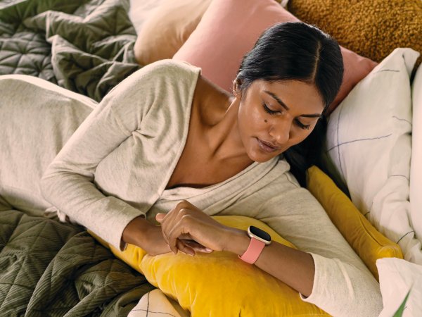 A woman in bed wearing a white long-sleeved shirt, looking at a pink Fitbit on her wrist.