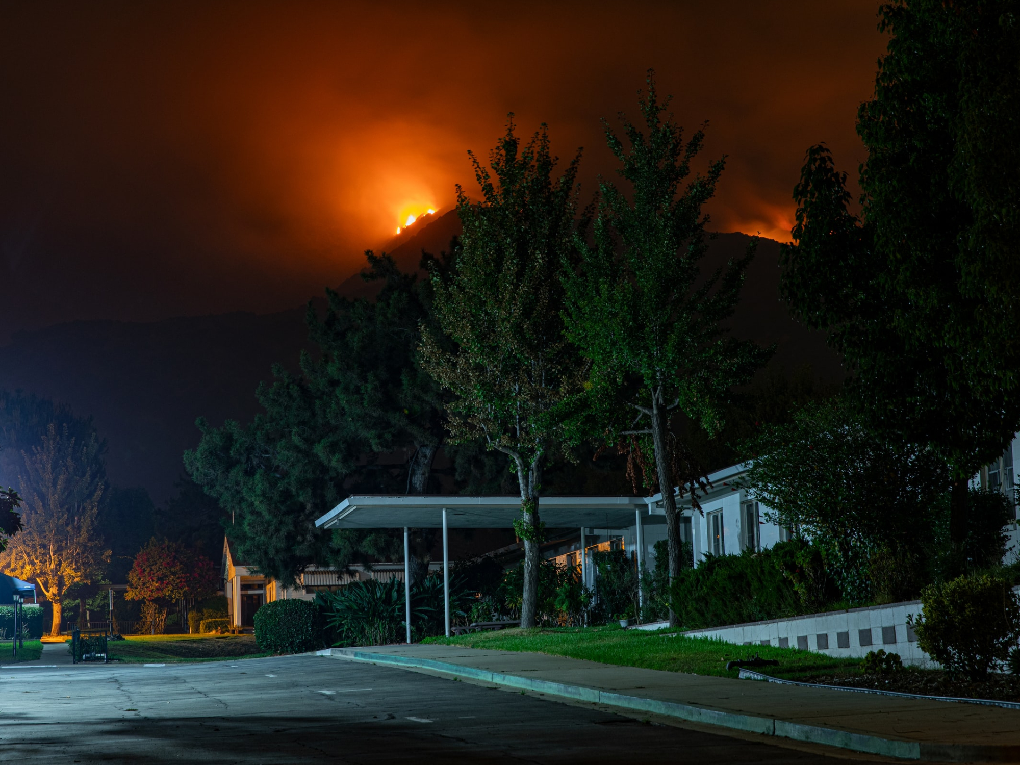 A large home in California being threatened by a wildfire on a distant mountain at night.