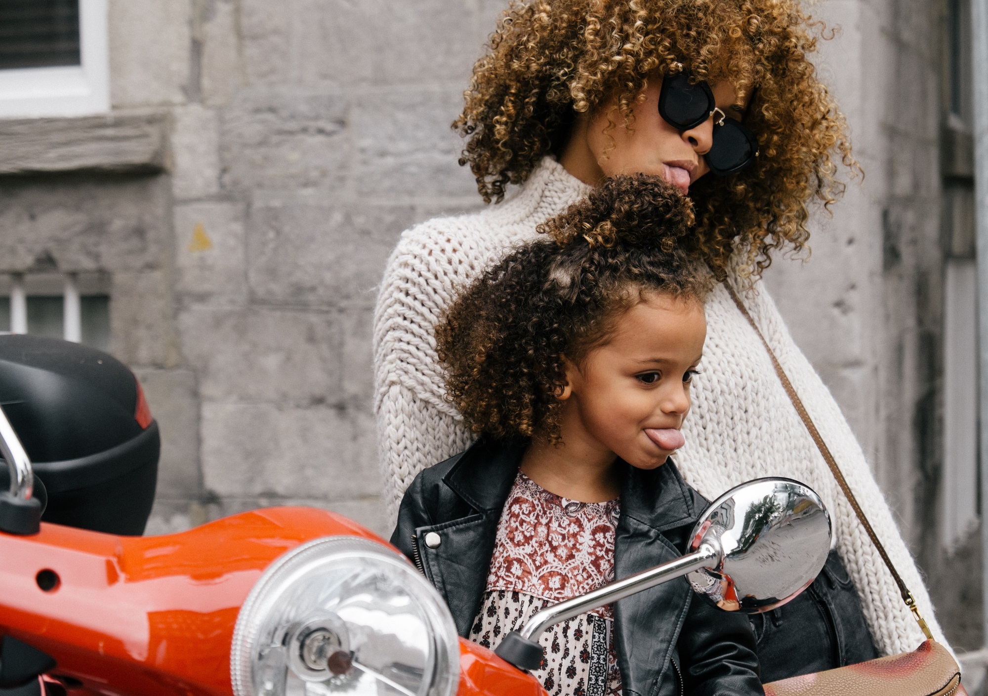 Gen Z and millennial kid and parent with curly hair and leather jackets sticking their tongues out in the mirror of a red motorcycle