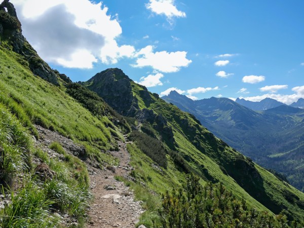 A rocky trail on the side of a steep, grassy mountain under a blue sky. It's the kind of hike you'd need to prepare for.