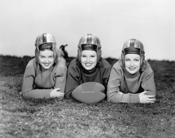 Three women in helmets leaning down on turf with a foam heart-shaped football in black and white