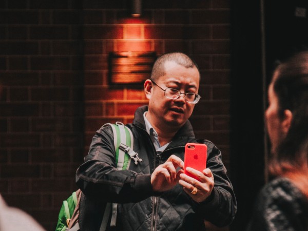 An Asian man with short hair and glasses wearing a black jacket and a green backpack while zooming in on something on the screen of his iPhone.
