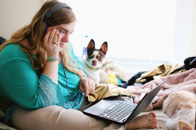 College students in headphones and teal shirt sitting on bed with laptop and small brown and white dog trying to get broadband access