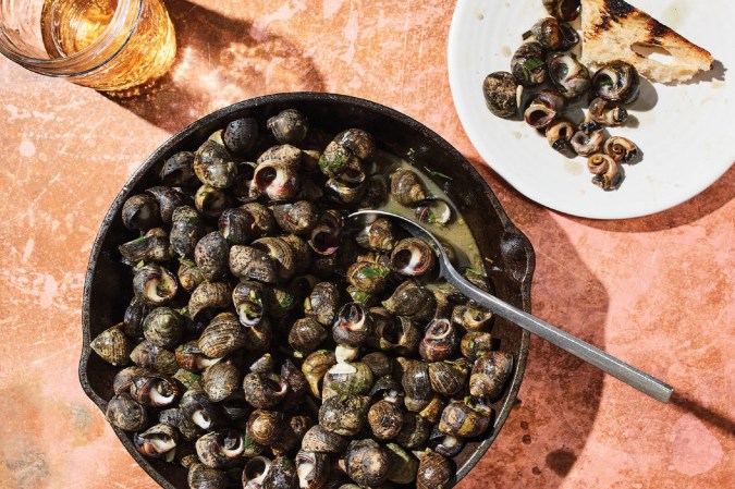 Periwinkles in a saucepan next to a small plate of bread and a glass of cider from an invasives-species menu at Juniper Bar and Restaurant in Burlington, Vermont