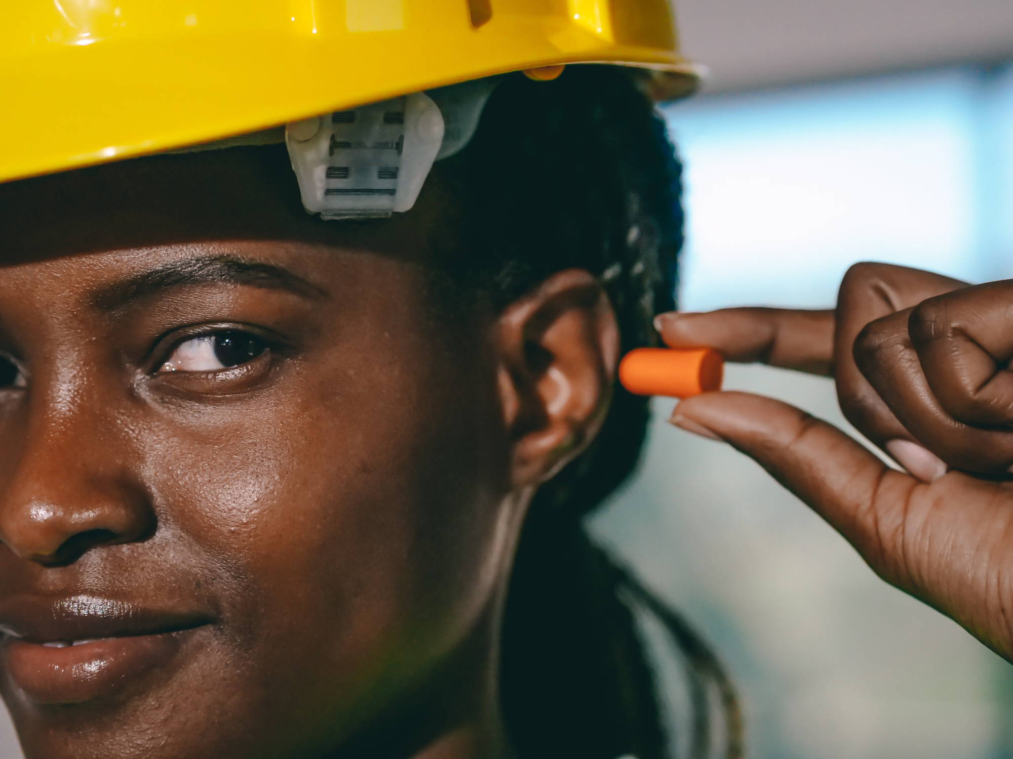 A Black woman wearing a yellow hard hat while inserting an orange foam ear plug into her ear.