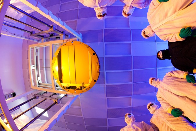 James Webb telescope primary mirror coated in gold seen from a fisheye view looking up with engineers in masks and protective gear looking down from a blue ceiling