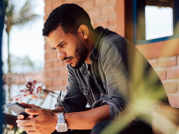 A man sitting on a bench outside a brick building looking intensely at his phone.